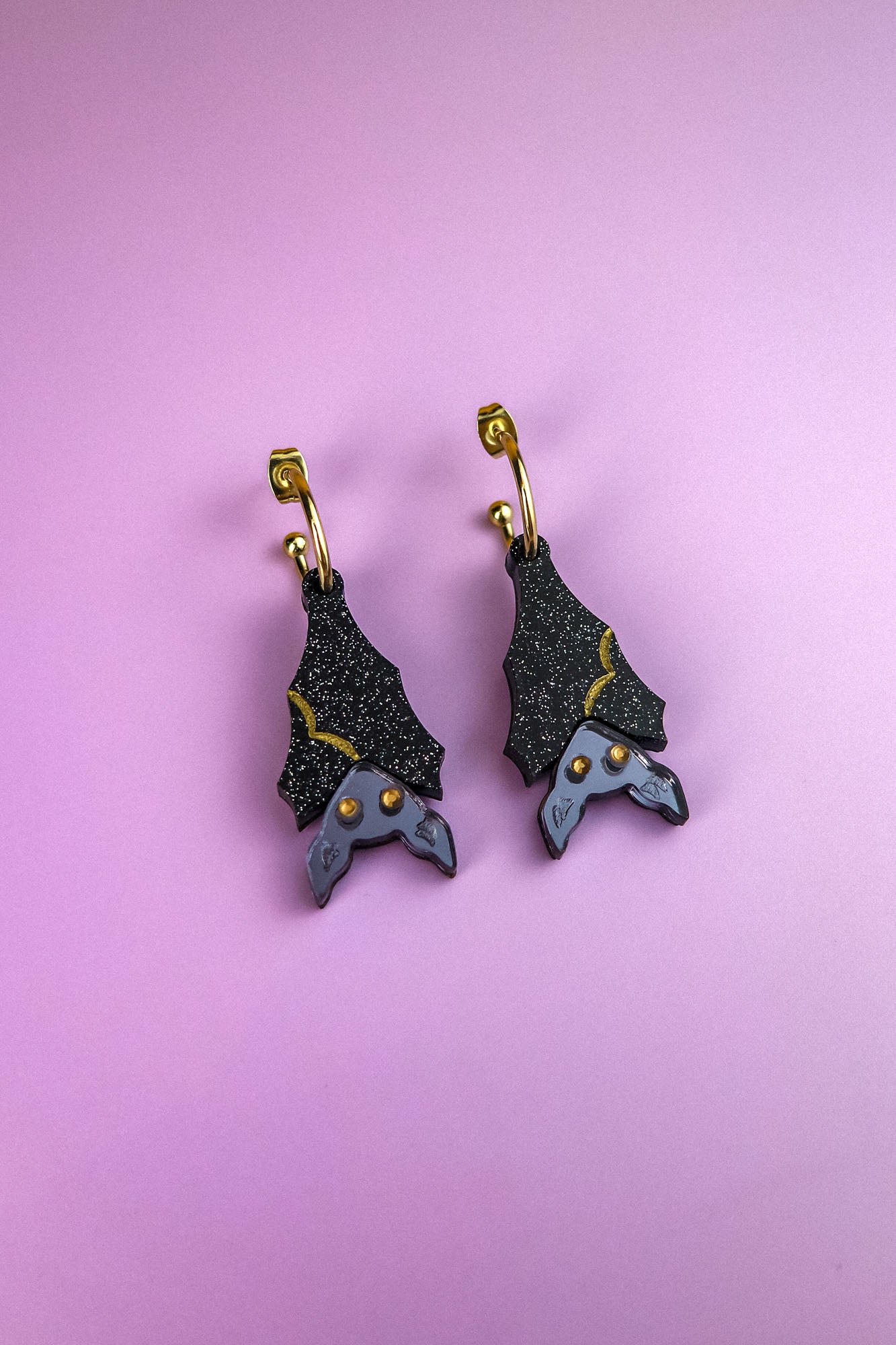 A pair of hand-painted bat-shaped earrings with black, glittery wings and gold accents, hanging from gold hoops, displayed on a pastel purple background. These allergy-free earrings combine unique design with comfortable wear.