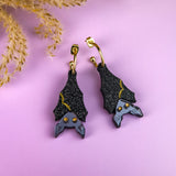 A pair of hand-painted, black bat-shaped earrings with gold accents and gray faces, displayed on a pastel purple surface with dried beige foliage in the corner.