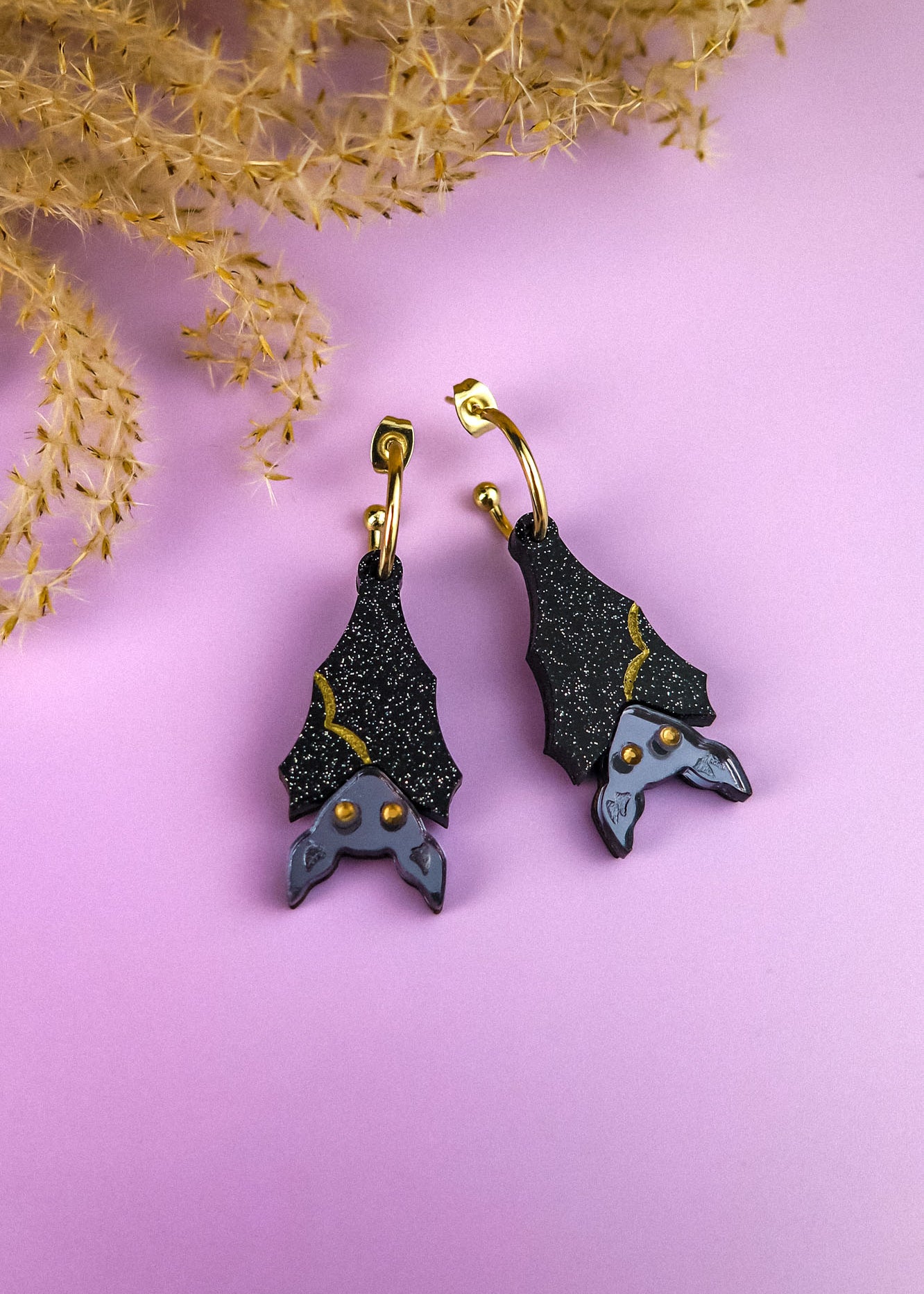 A pair of hand-painted, black bat-shaped earrings with gold accents and gray faces, displayed on a pastel purple surface with dried beige foliage in the corner.