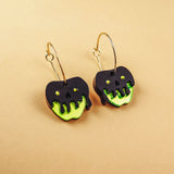 A pair of limited edition Poisoned Apple Earrings shaped like apples with neon green bottoms and black, drippy tops resembling poison, hanging from gold hoops, displayed on a beige background.