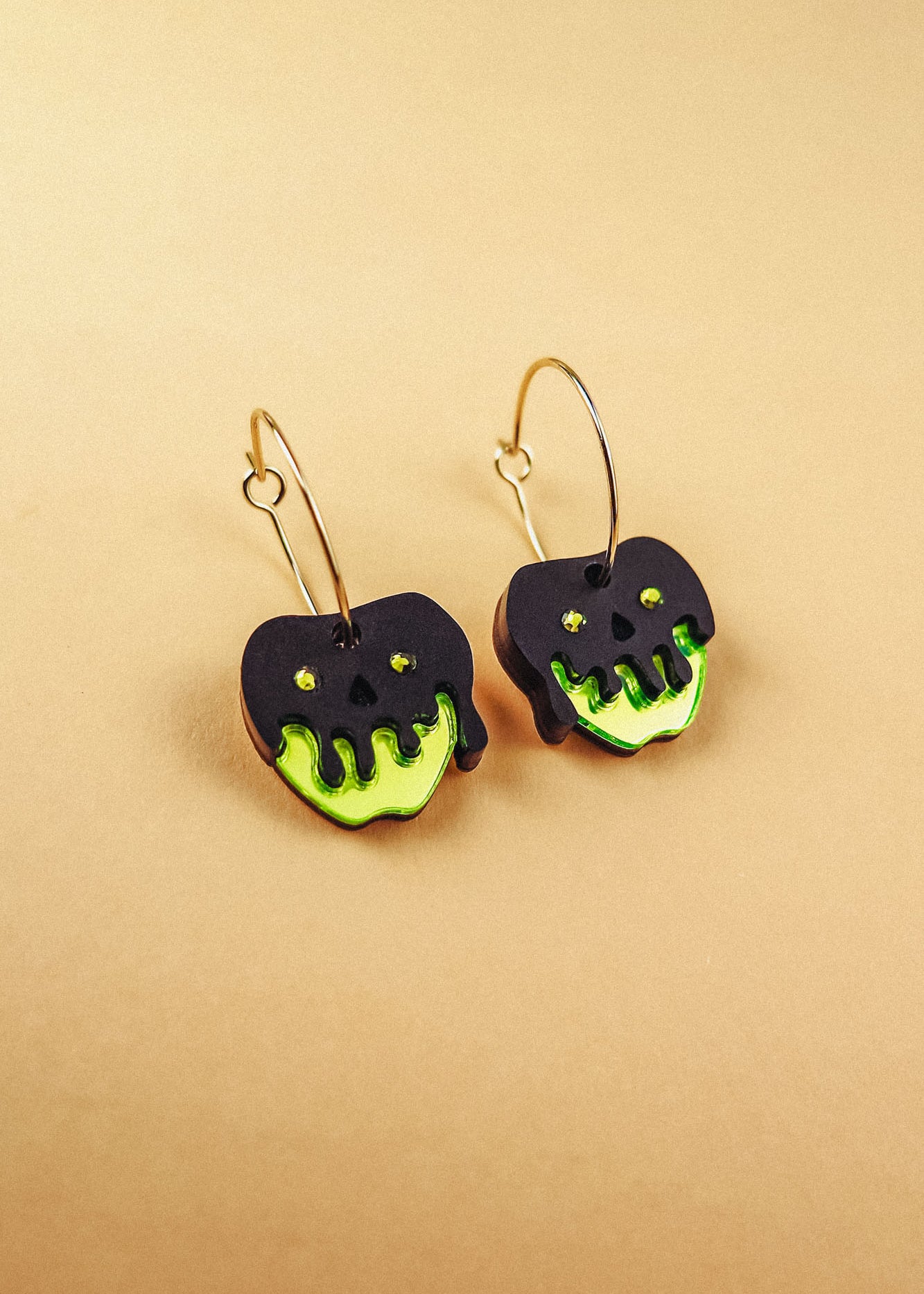 A pair of limited edition Poisoned Apple Earrings shaped like apples with neon green bottoms and black, drippy tops resembling poison, hanging from gold hoops, displayed on a beige background.