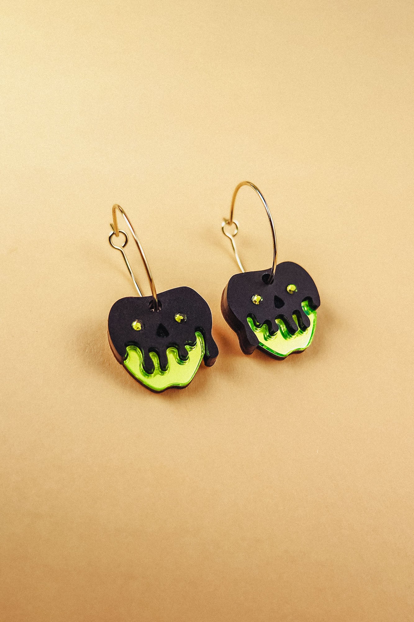 A pair of limited edition Poisoned Apple Earrings shaped like apples with neon green bottoms and black, drippy tops resembling poison, hanging from gold hoops, displayed on a beige background.
