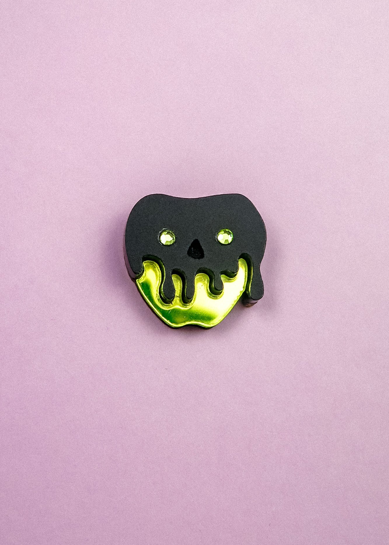 A pin shaped like an apple with a shiny yellow bottom and a black, drippy skull design on top, this Poisoned Apple Badge is perfect for Halloween lovers. Set against a plain light purple background—grab yours now, as stock is limited!.