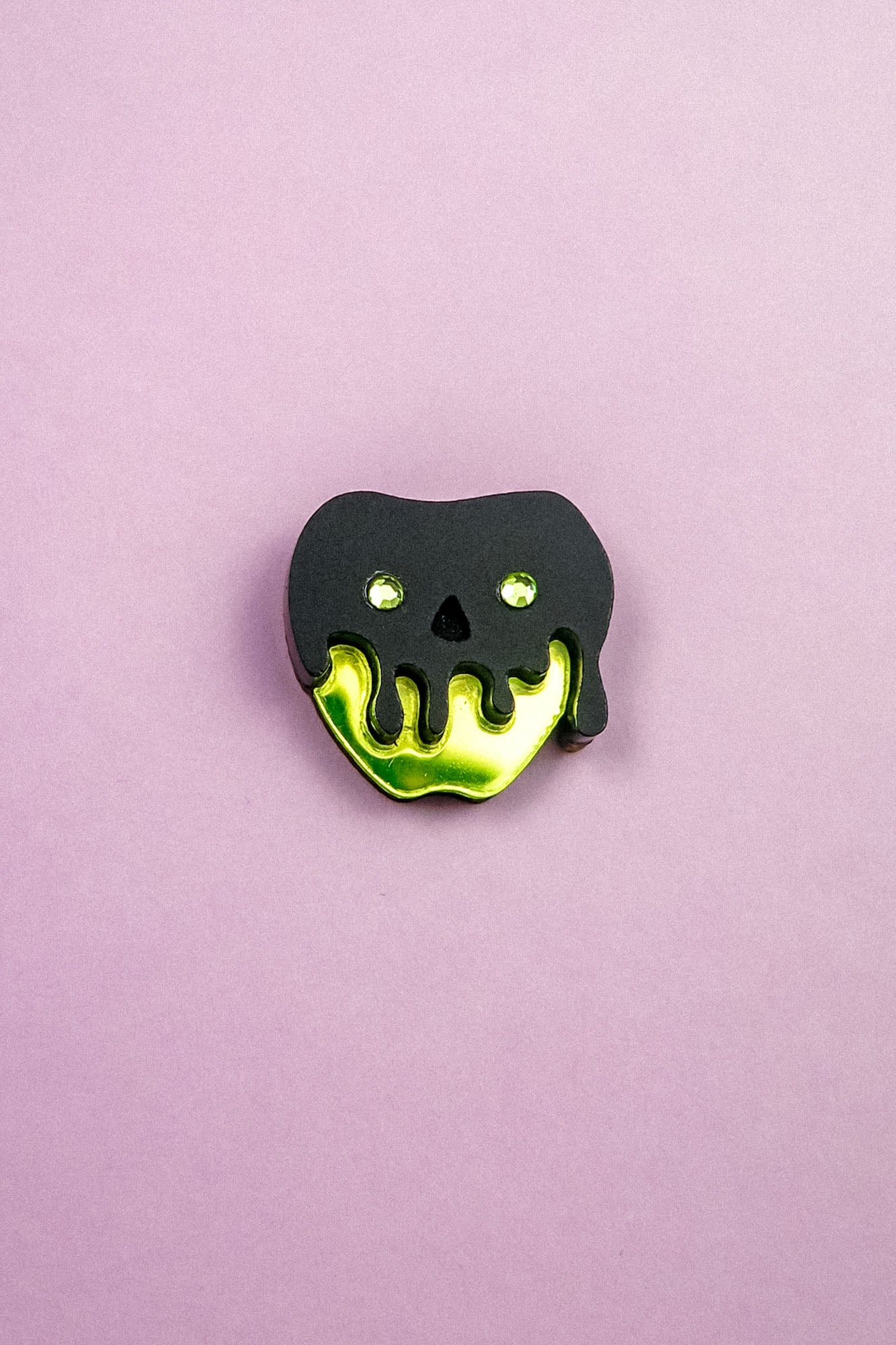 A pin shaped like an apple with a shiny yellow bottom and a black, drippy skull design on top, this Poisoned Apple Badge is perfect for Halloween lovers. Set against a plain light purple background—grab yours now, as stock is limited!.