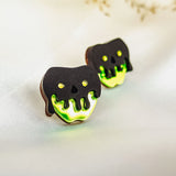 Two cookies decorated to look like poison apples, with black and neon green icing resembling a dripping skull, are placed on a light fabric surface. Their handcrafted details perfectly capture a Halloween look against the blurred floral background.