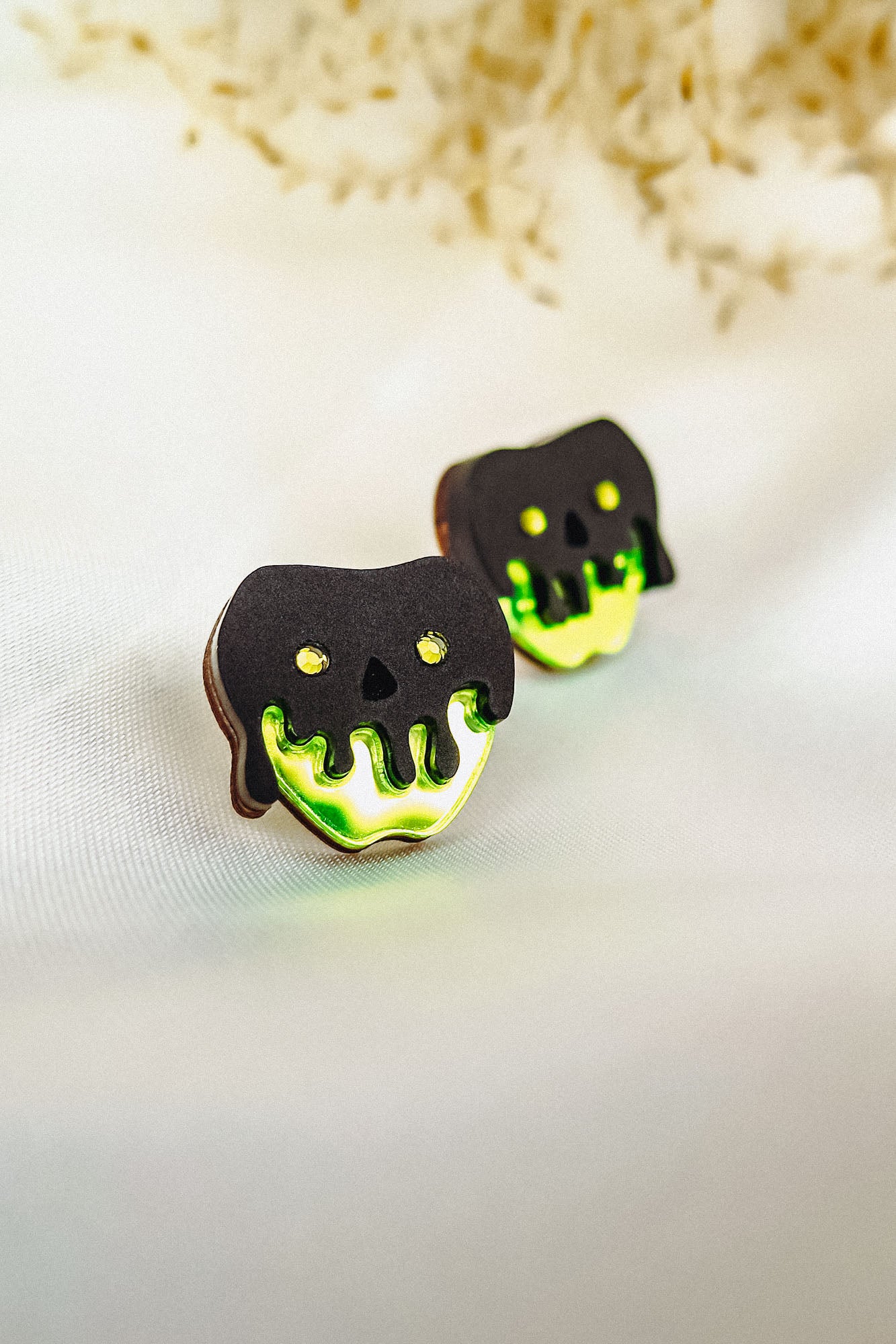 Two cookies decorated to look like poison apples, with black and neon green icing resembling a dripping skull, are placed on a light fabric surface. Their handcrafted details perfectly capture a Halloween look against the blurred floral background.
