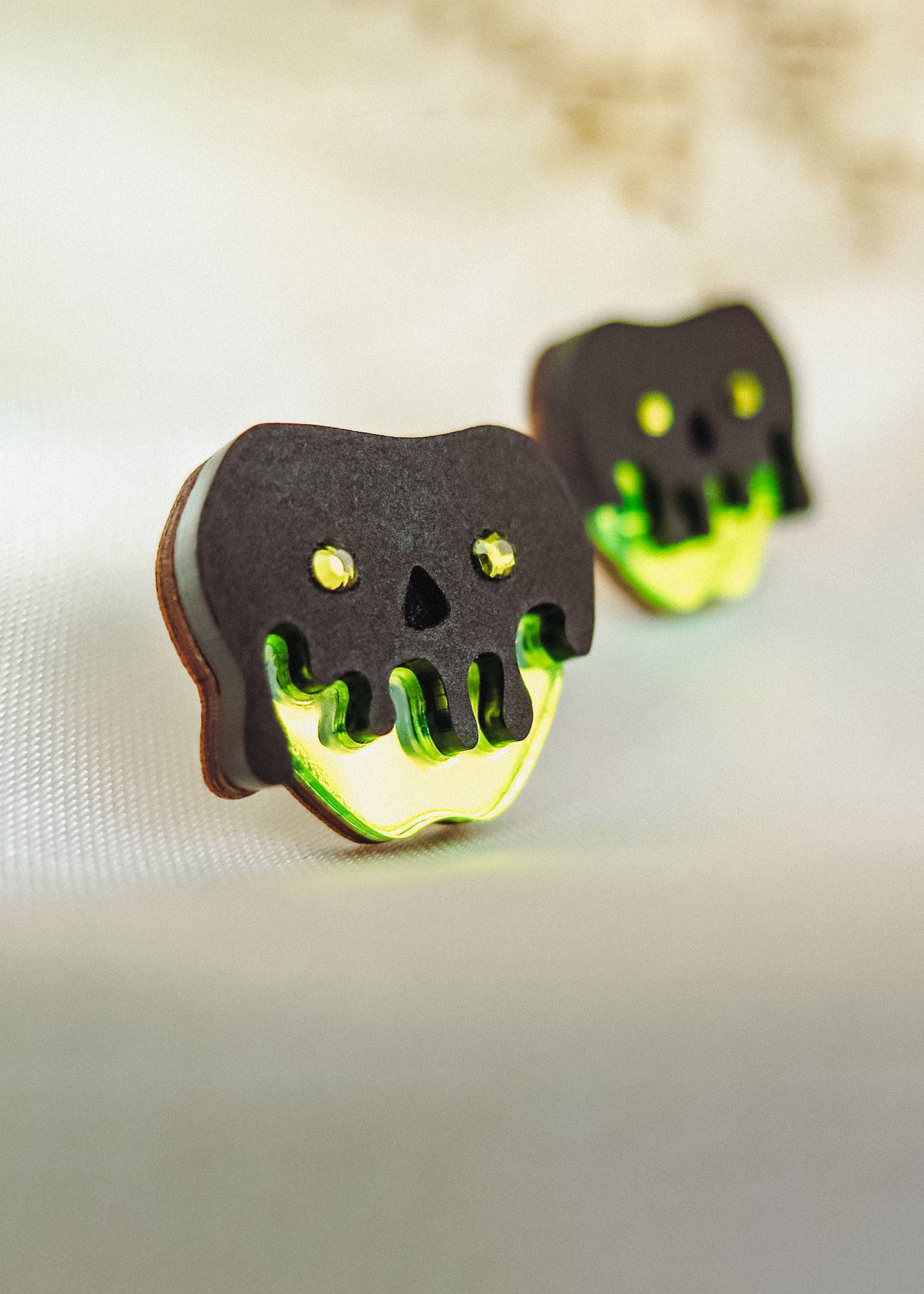 Two handcrafted cookies shaped like skulls with black tops and neon green dripping icing, featuring yellow eyes and nose holes, deliver a unique Halloween look as they rest on a light fabric surface.