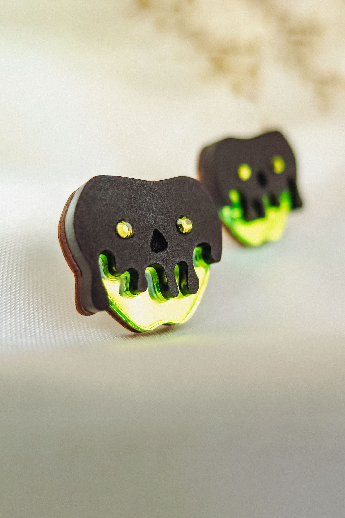 Two handcrafted cookies shaped like skulls with black tops and neon green dripping icing, featuring yellow eyes and nose holes, deliver a unique Halloween look as they rest on a light fabric surface.