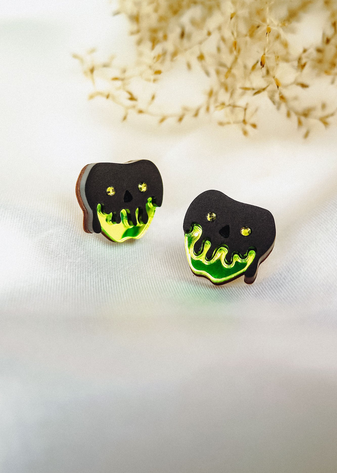 A pair of handcrafted earrings shaped like black skulls with glowing green drips at the bottom, perfect for a bold Halloween look, displayed on a white surface with dried grass in the background.