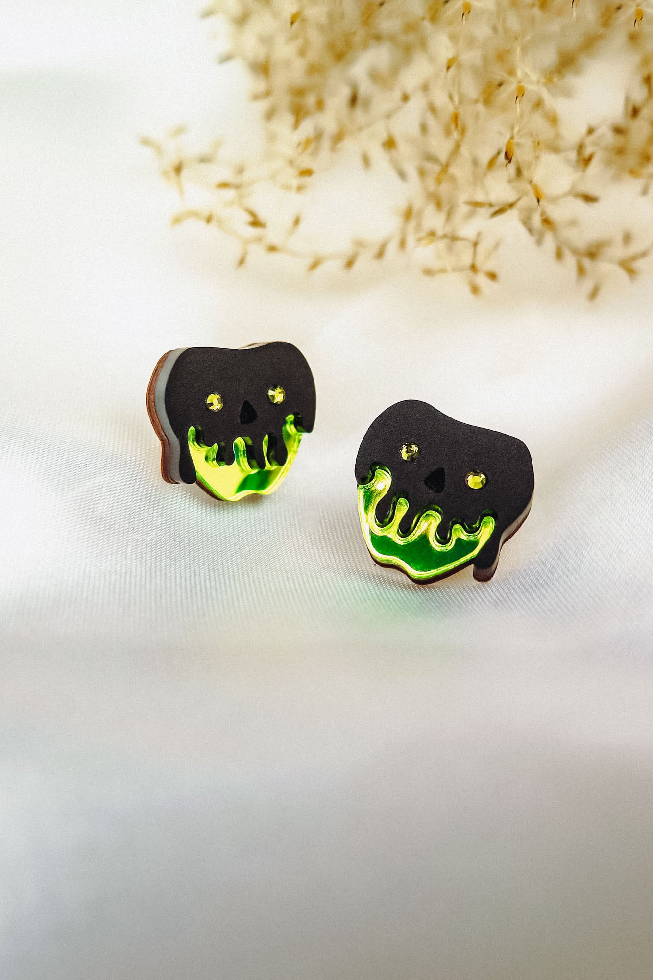 A pair of handcrafted earrings shaped like black skulls with glowing green drips at the bottom, perfect for a bold Halloween look, displayed on a white surface with dried grass in the background.
