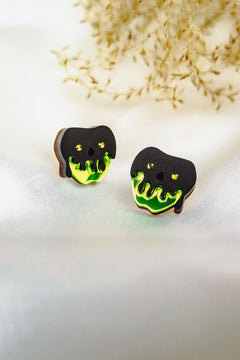 A pair of handcrafted earrings shaped like black skulls with glowing green drips at the bottom, perfect for a bold Halloween look, displayed on a white surface with dried grass in the background.