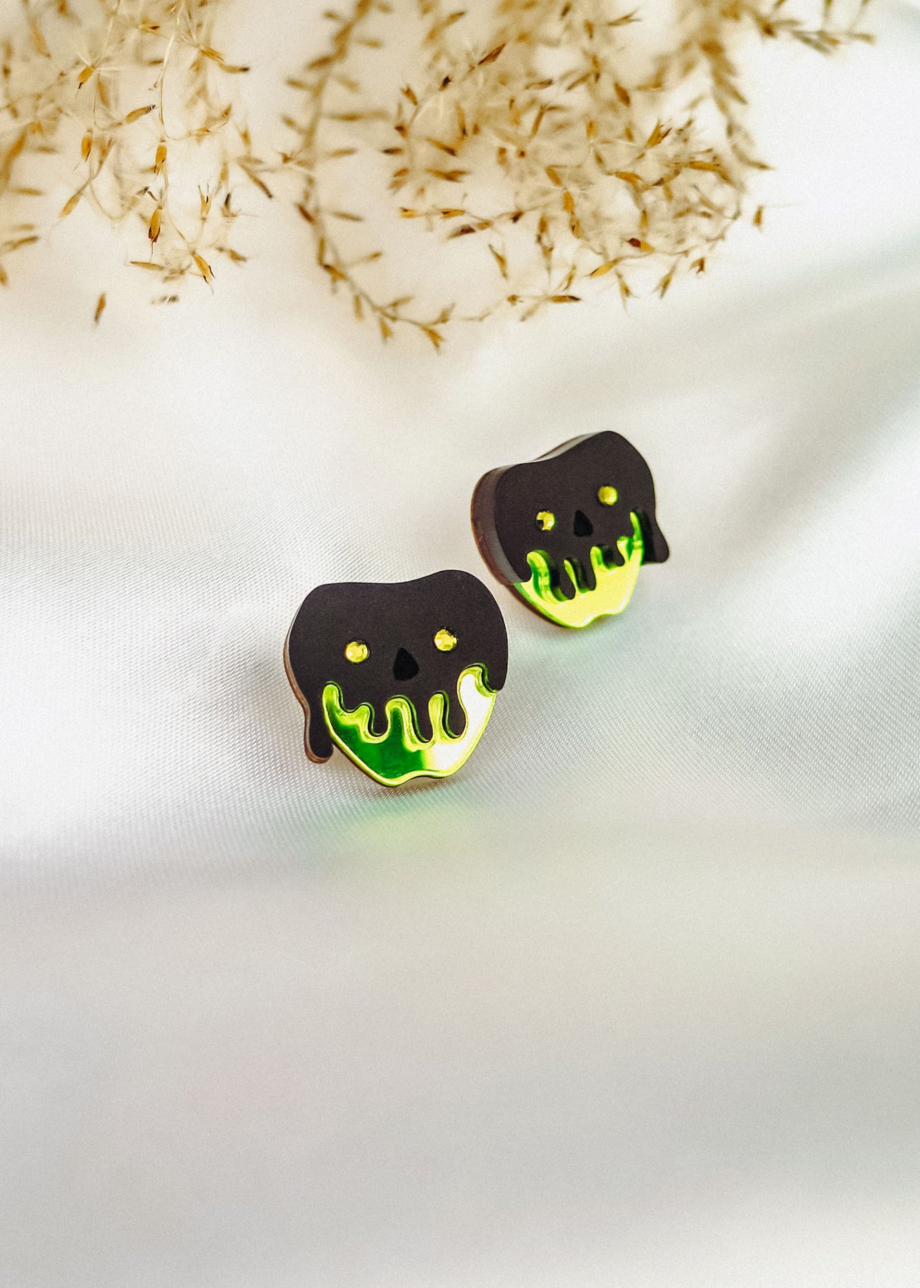 Two cookies shaped like apples with black and neon green dripping icing, resembling a poison apple, showcase a handcrafted Halloween look as they sit on a white fabric surface with delicate dried grass in the background.
