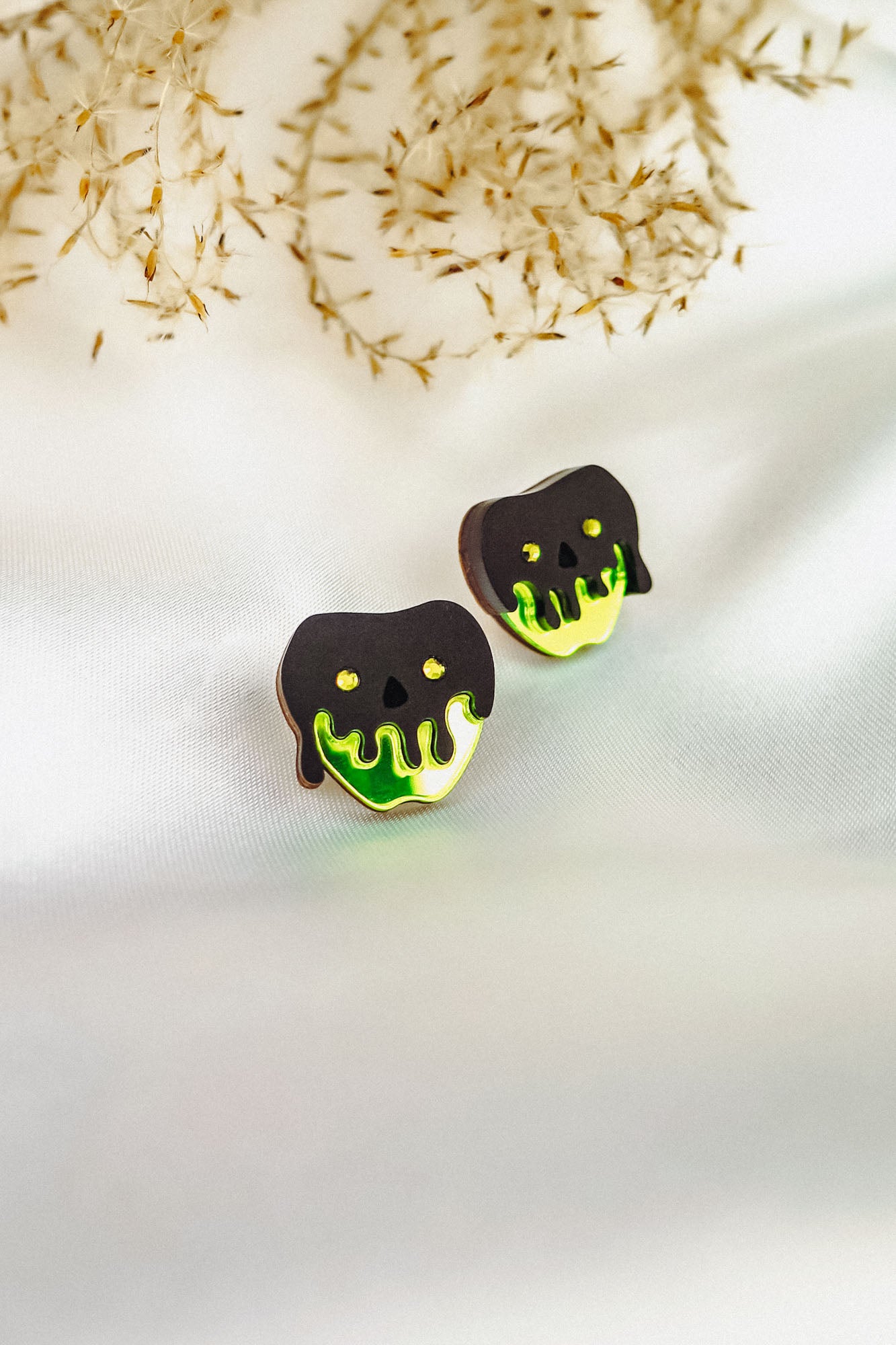 Two cookies shaped like apples with black and neon green dripping icing, resembling a poison apple, showcase a handcrafted Halloween look as they sit on a white fabric surface with delicate dried grass in the background.