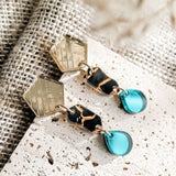 A pair of stylish, handcrafted tear-drop earrings featuring gold hexagonal studs, black and gold patterned middle segments, and translucent blue dangles, displayed on textured fabric and stone surfaces.