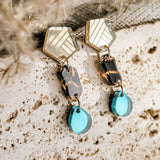 A pair of eco-friendly, handcrafted geometric dangle earrings with gold hexagon tops, marbled brown and black middle links, and translucent blue tear-drop earrings, displayed on a textured beige stone surface.