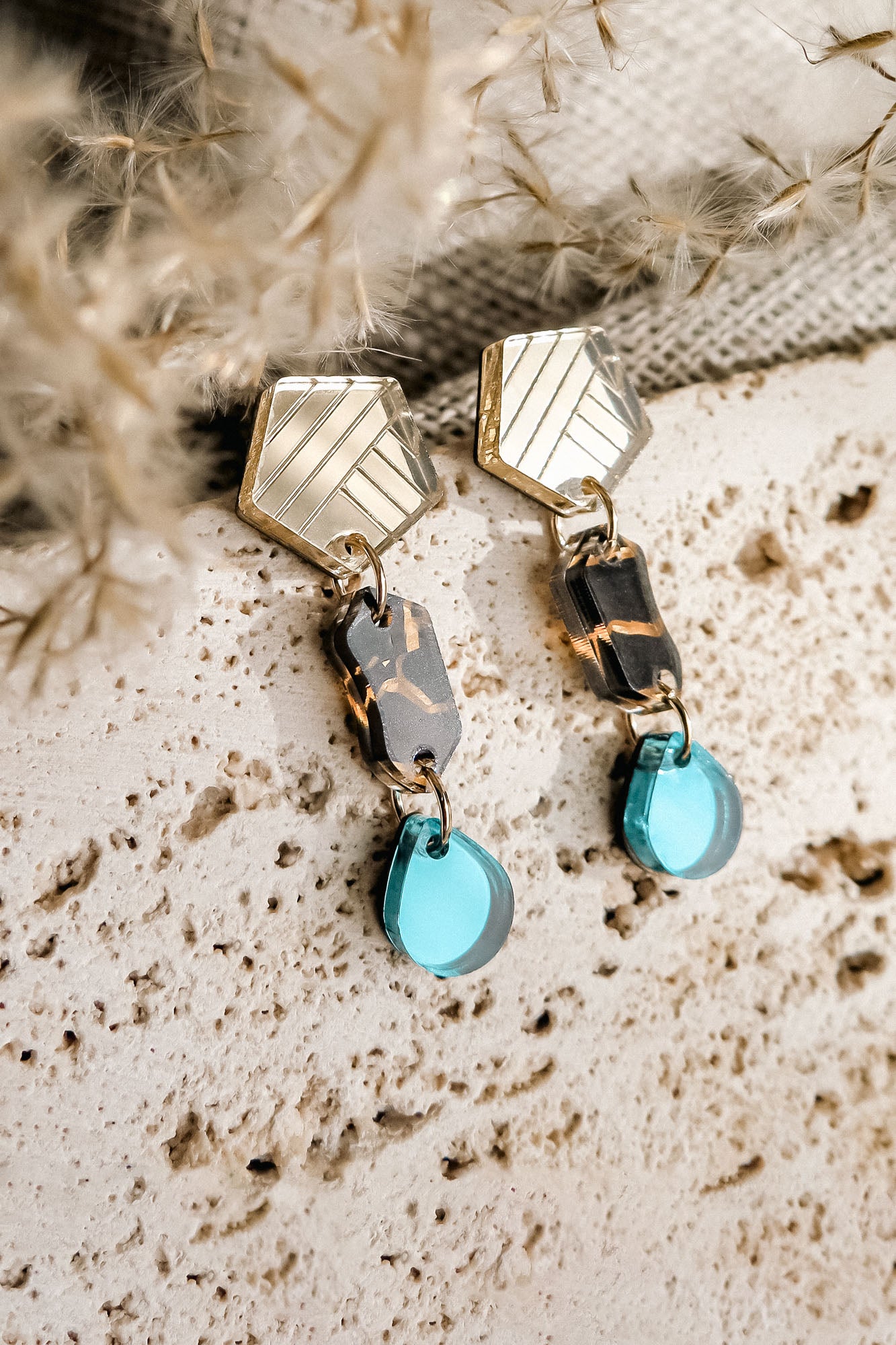A pair of eco-friendly, handcrafted geometric dangle earrings with gold hexagon tops, marbled brown and black middle links, and translucent blue tear-drop earrings, displayed on a textured beige stone surface.