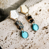A pair of handcrafted geometric drop earrings with gold, tortoiseshell, and turquoise blue elements rest on a textured stone surface with a neutral fabric and dried plants in the background.