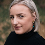 A woman with short, straight blonde hair and handcrafted gold earrings looks at the camera with a slight smile, wearing a black turtleneck sweater. The blurred background features eco-friendly greenery and warm brown tones.