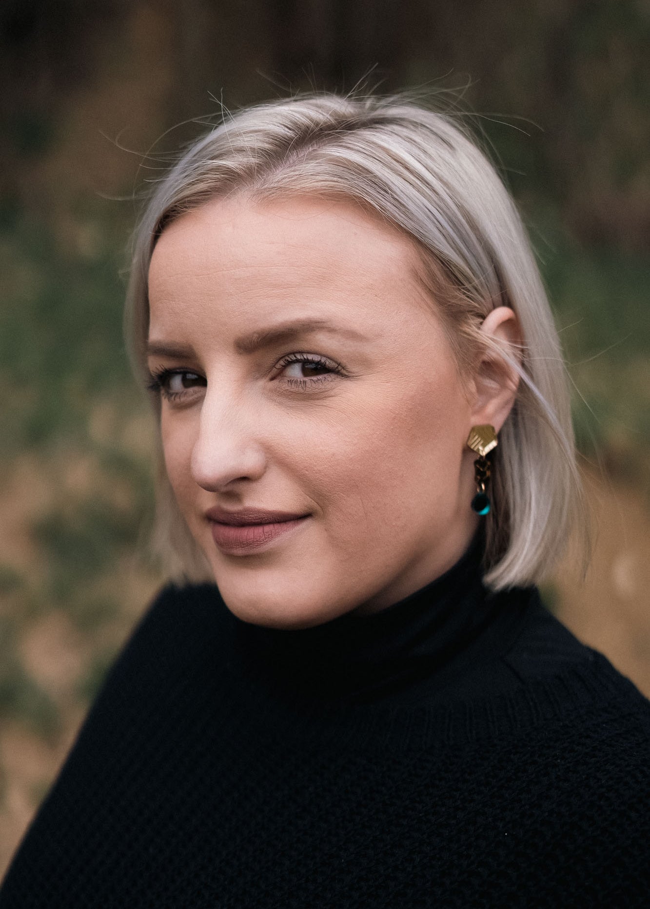 A woman with short, straight blonde hair and handcrafted gold earrings looks at the camera with a slight smile, wearing a black turtleneck sweater. The blurred background features eco-friendly greenery and warm brown tones.