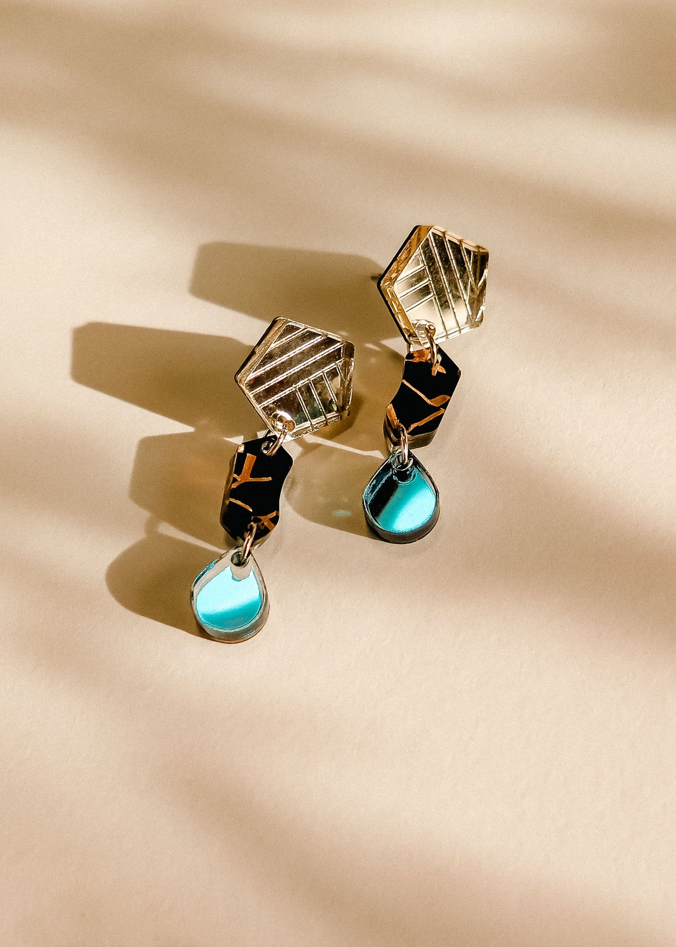 A pair of handcrafted geometric earrings with gold angular tops, black middle sections, and blue tear-drop pendants, displayed on a light beige surface with soft shadow patterns.
