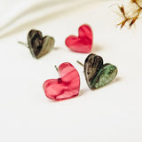 Four hypoallergenic, heart-shaped stud earrings—two in glossy pink and two in marbled green and black—are displayed on a white surface with dried plants, capturing minimalist charm.