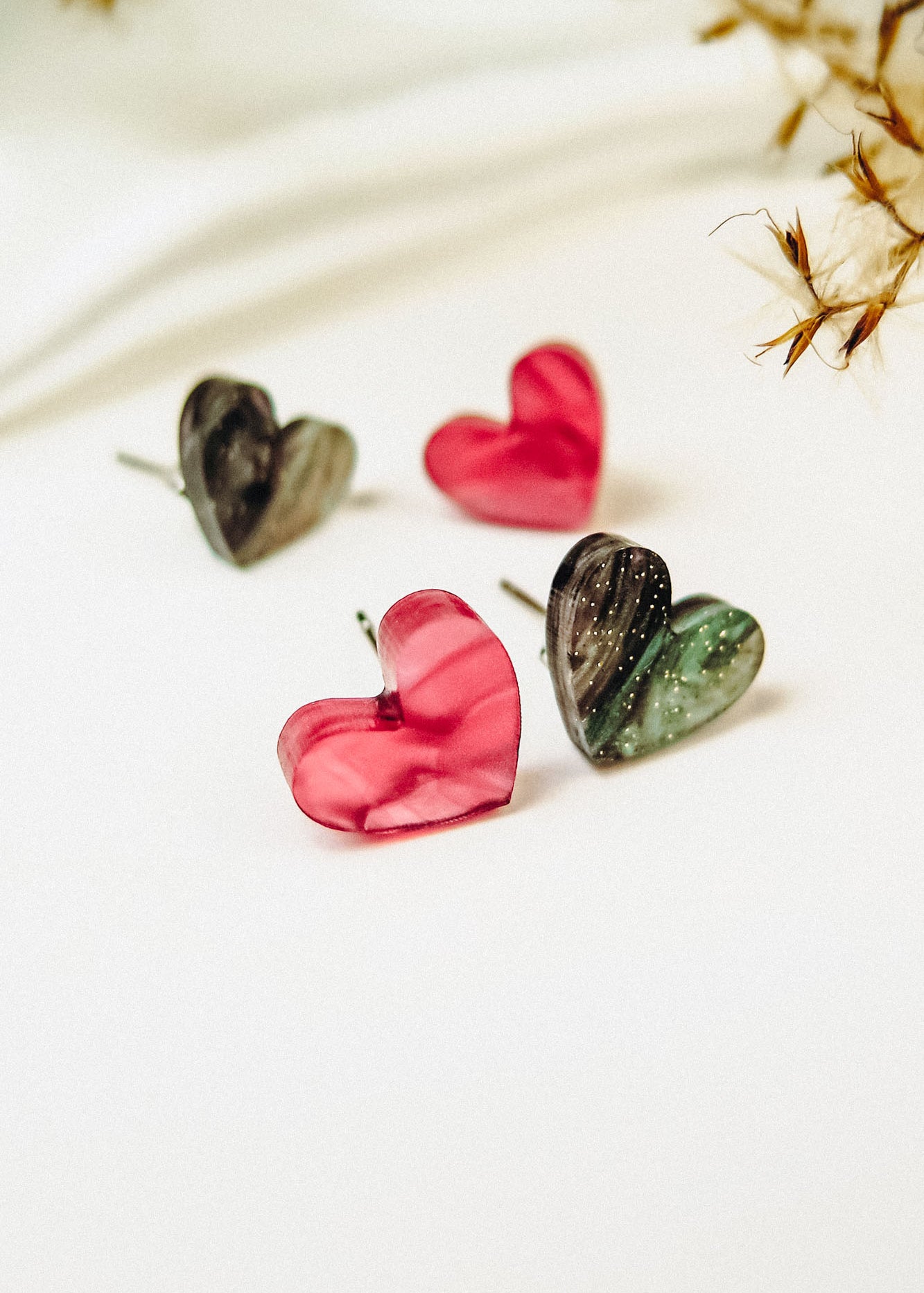 Four hypoallergenic, heart-shaped stud earrings—two in glossy pink and two in marbled green and black—are displayed on a white surface with dried plants, capturing minimalist charm.