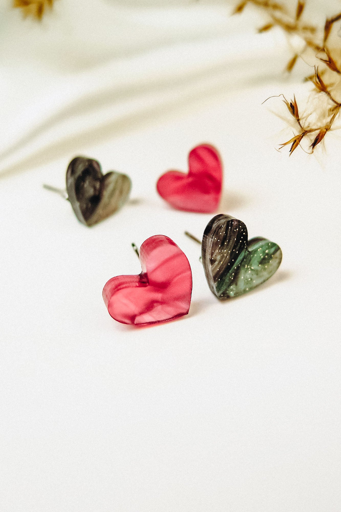 Four hypoallergenic, heart-shaped stud earrings—two in glossy pink and two in marbled green and black—are displayed on a white surface with dried plants, capturing minimalist charm.