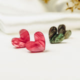 A pair of pink Tiny Heart Studs and dark green heart-shaped earrings are displayed on a white surface, radiating minimalist charm against a blurred white and yellow floral background. Hypoallergenic design ensures comfort for sensitive ears.