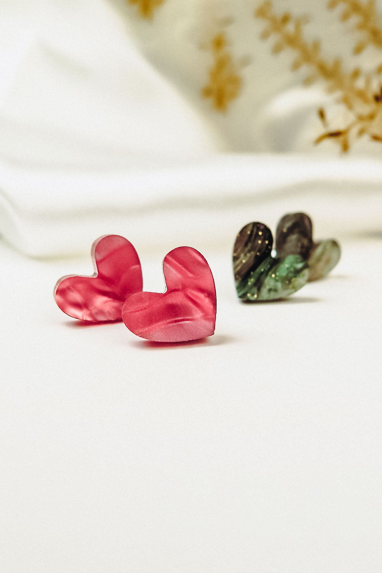 A pair of pink Tiny Heart Studs and dark green heart-shaped earrings are displayed on a white surface, radiating minimalist charm against a blurred white and yellow floral background. Hypoallergenic design ensures comfort for sensitive ears.