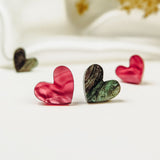 Four heart-shaped objects, resembling Tiny Heart Studs in pink and green with a marbled texture, are arranged on a white surface against a blurred white and gold background, radiating minimalist charm.