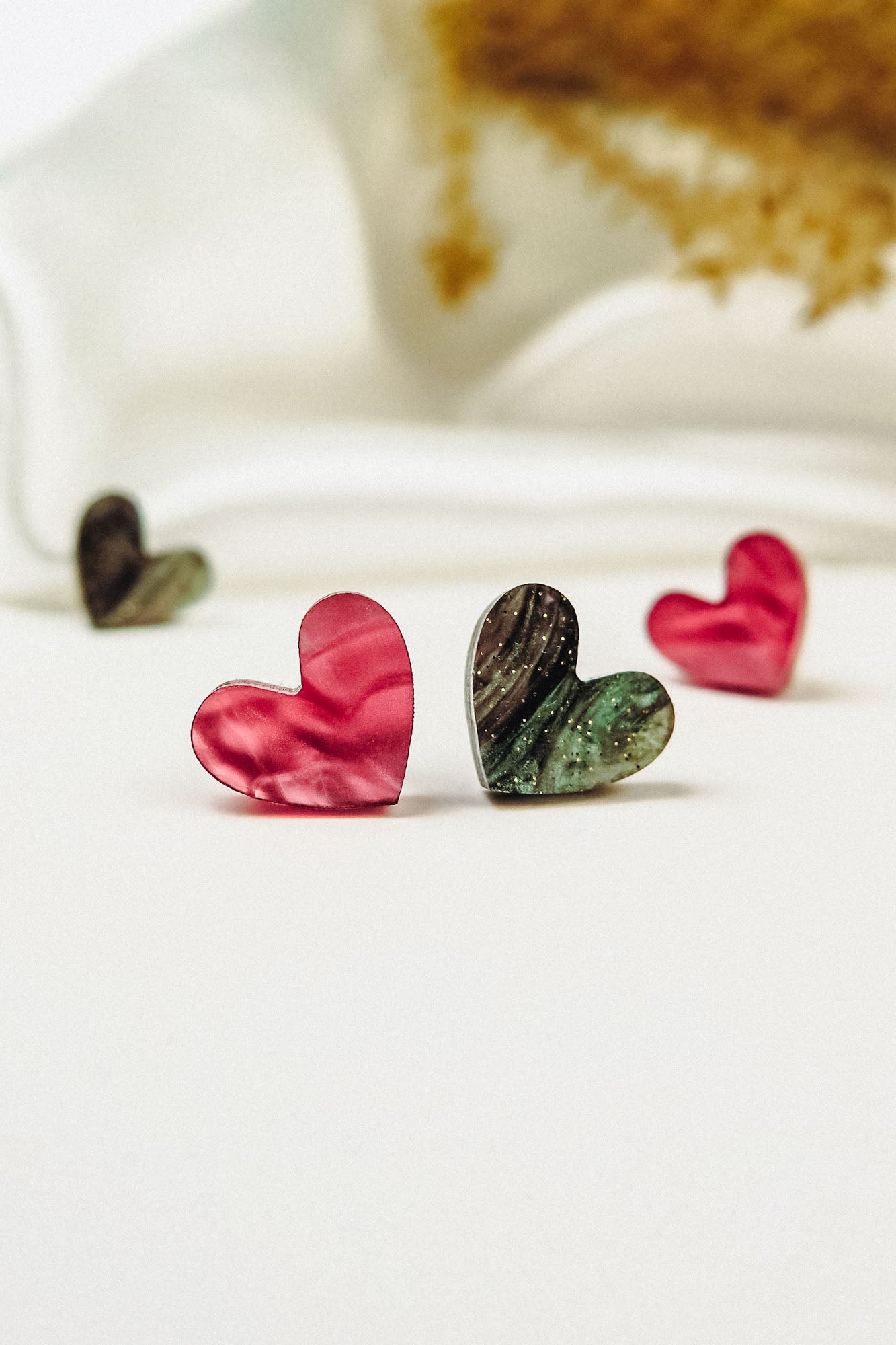 Four heart-shaped objects, resembling Tiny Heart Studs in pink and green with a marbled texture, are arranged on a white surface against a blurred white and gold background, radiating minimalist charm.
