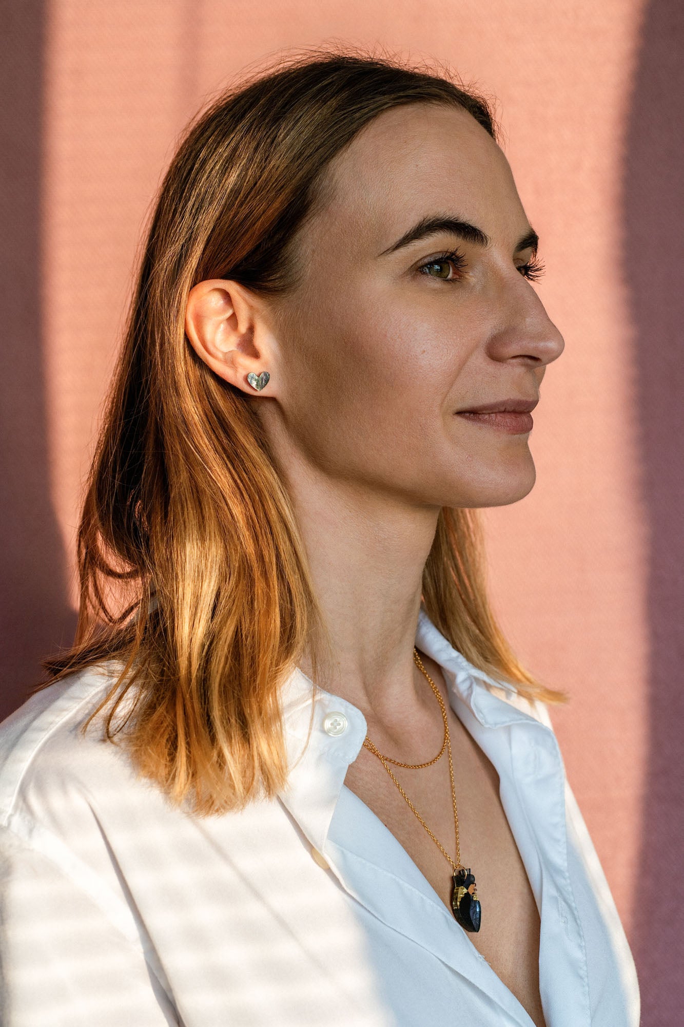 A woman with straight, shoulder-length light brown hair, wearing a white shirt, layered gold necklaces, and tiny heart studs, stands in front of a pink background, looking to the right with a calm expression.