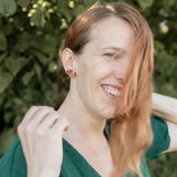 A woman with long, light brown hair wearing a green top smiles outdoors, tiny heart studs adding minimalist charm. She stands in front of leafy green foliage as sunlight filters through the trees, with some hair falling across her face.