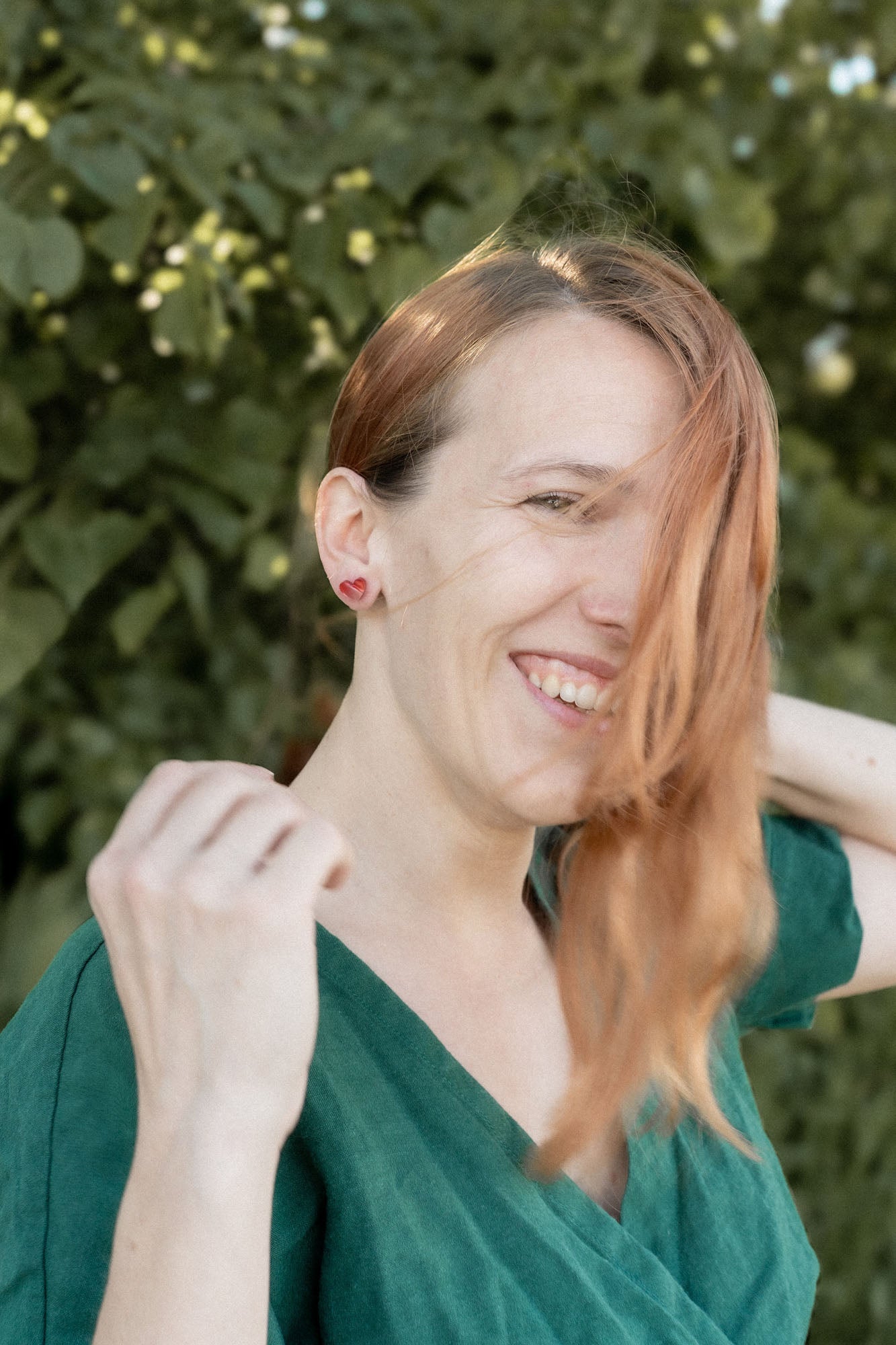 A woman with long, light brown hair wearing a green top smiles outdoors, tiny heart studs adding minimalist charm. She stands in front of leafy green foliage as sunlight filters through the trees, with some hair falling across her face.