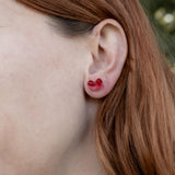 Close-up of a person with long reddish-brown hair wearing a red Tiny Heart Stud in their left ear, highlighting its minimalist charm. The background is blurred greenery.