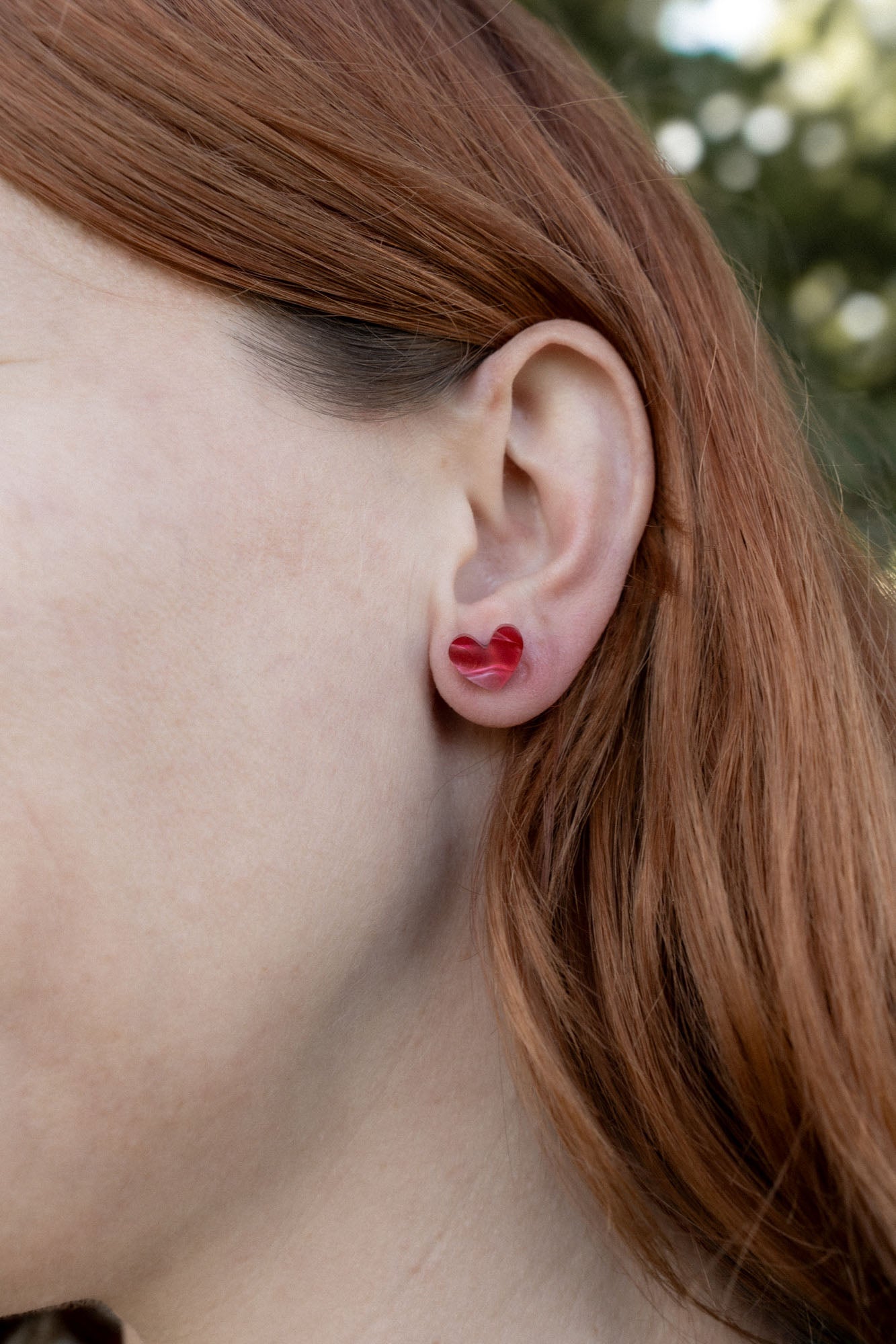 Close-up of a person with long reddish-brown hair wearing a red Tiny Heart Stud in their left ear, highlighting its minimalist charm. The background is blurred greenery.