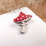 A handcrafted, eco-friendly Toadstool Mushroom Pin, shaped and painted in red and white, sits on a white surface with dried grass or flowers softly blurred in the background.
