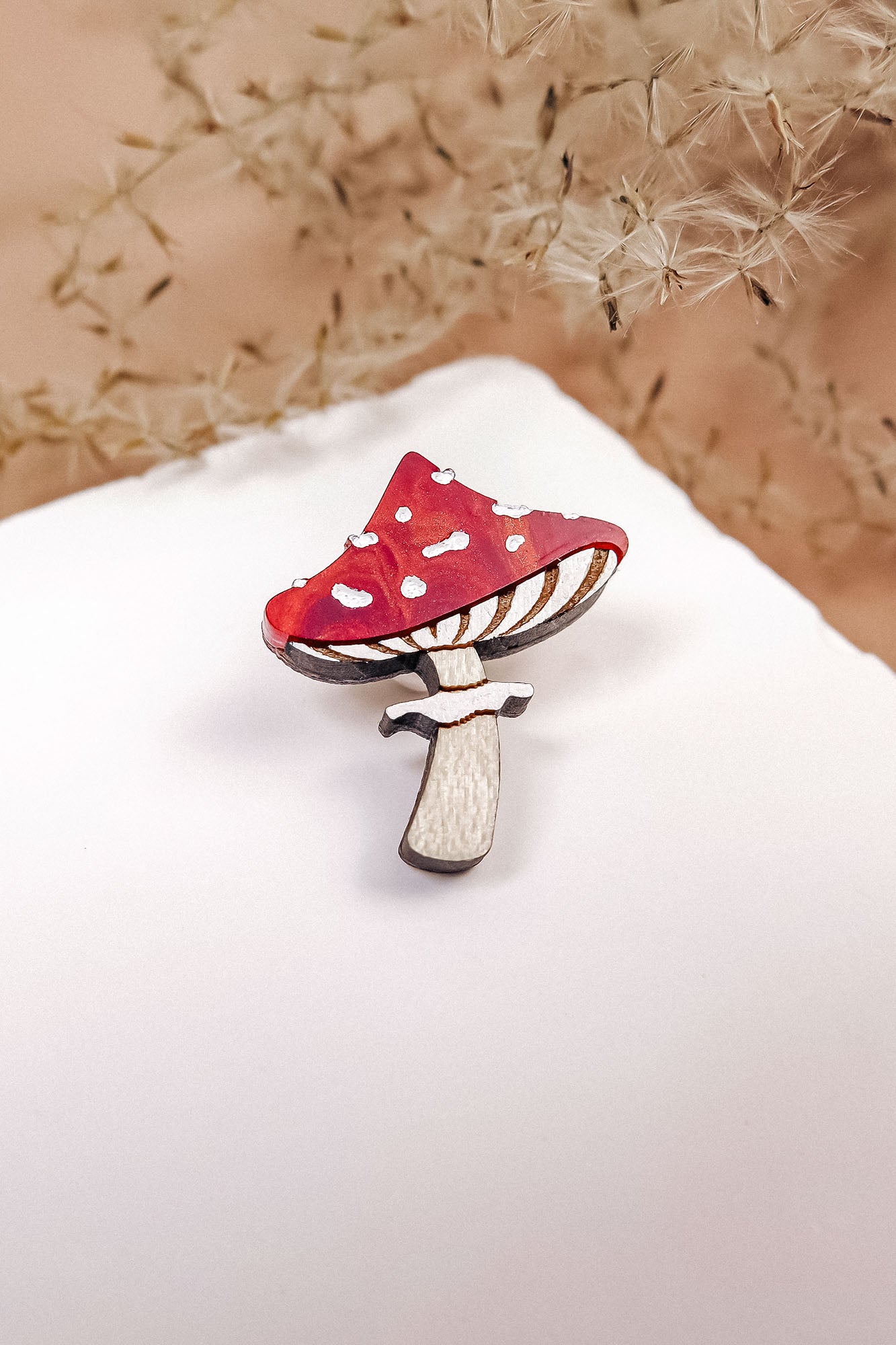 A handcrafted, eco-friendly Toadstool Mushroom Pin, shaped and painted in red and white, sits on a white surface with dried grass or flowers softly blurred in the background.