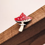 A small, handcrafted Toadstool Mushroom Pin shaped like a red and white spotted mushroom is attached to a wooden surface.