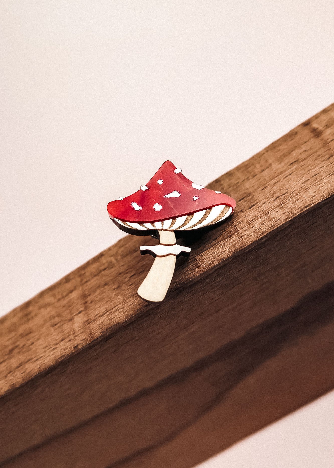 A small, handcrafted Toadstool Mushroom Pin shaped like a red and white spotted mushroom is attached to a wooden surface.