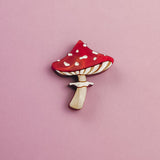 A handcrafted, eco-friendly Toadstool Mushroom Pin, shaped and painted like a red and white mushroom, rests on a light pink background.