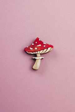 A handcrafted, eco-friendly Toadstool Mushroom Pin, shaped and painted like a red and white mushroom, rests on a light pink background.