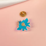 A blue, abstract-shaped Tooth Fairy Pin with a gold clasp backing rests on a soft pink surface, evoking childhood milestones, while a white fabric is draped in the top corner.
