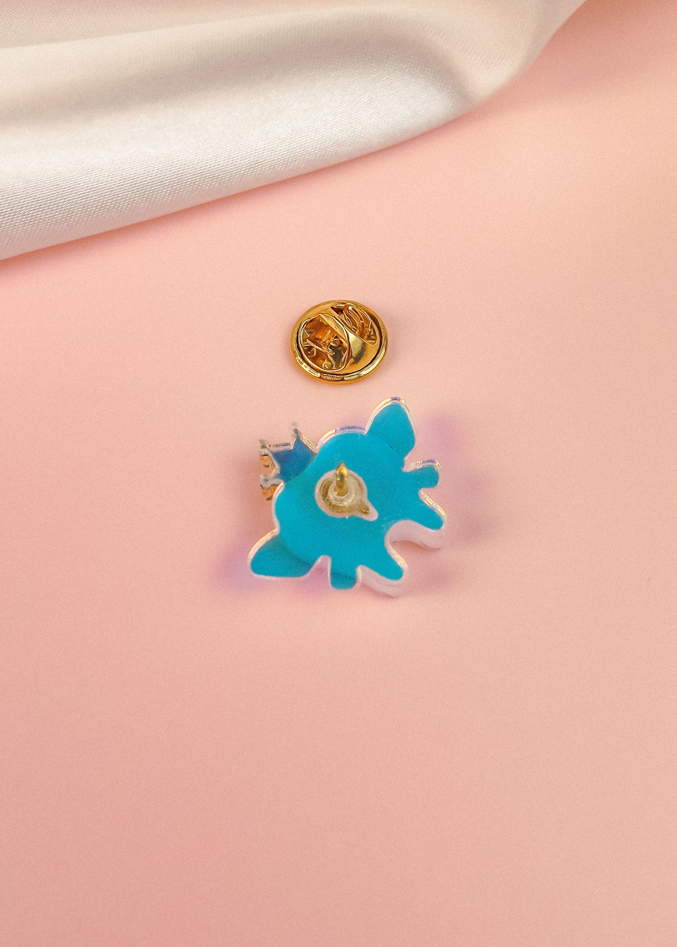 A blue, abstract-shaped Tooth Fairy Pin with a gold clasp backing rests on a soft pink surface, evoking childhood milestones, while a white fabric is draped in the top corner.