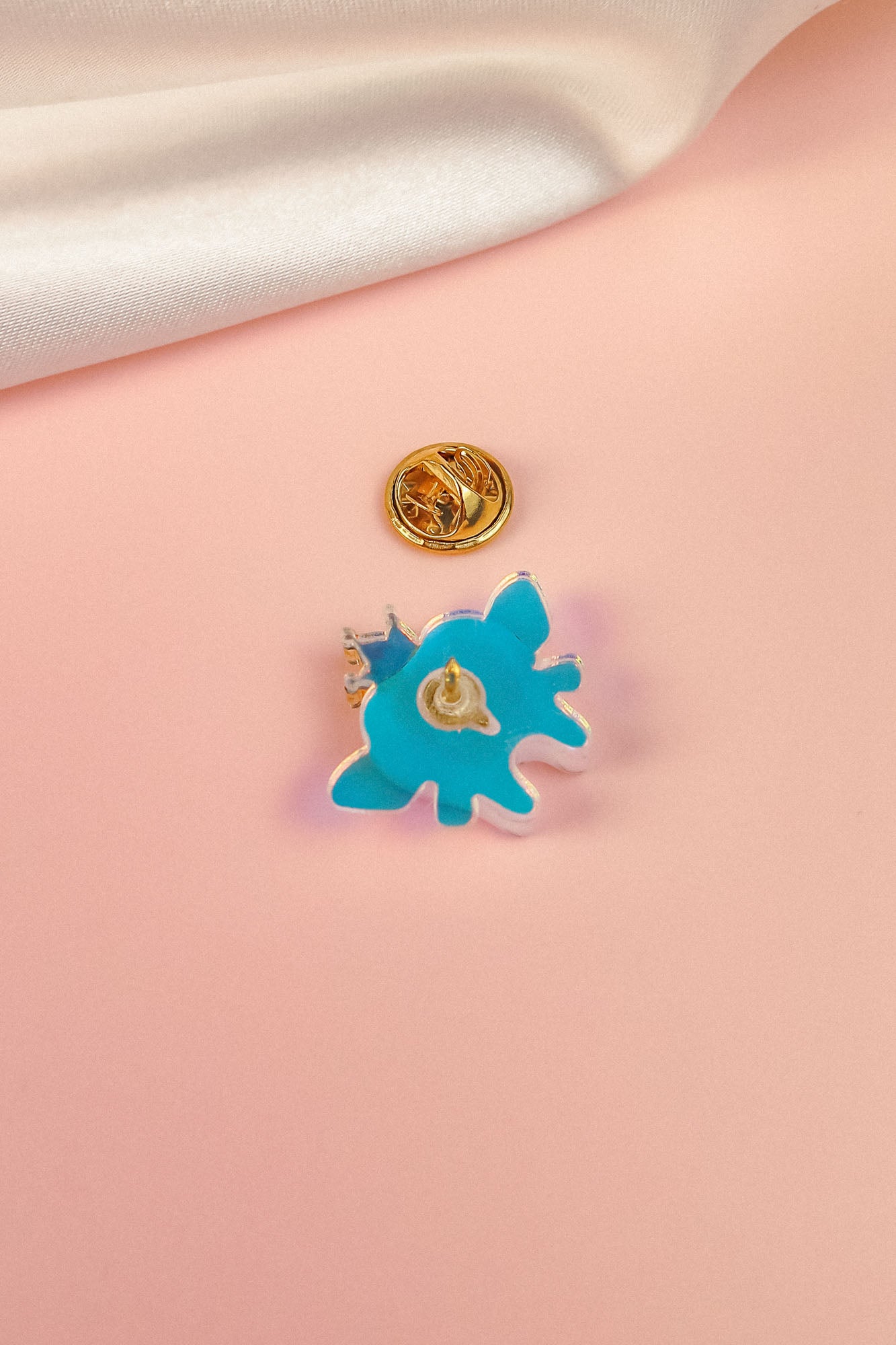 A blue, abstract-shaped Tooth Fairy Pin with a gold clasp backing rests on a soft pink surface, evoking childhood milestones, while a white fabric is draped in the top corner.