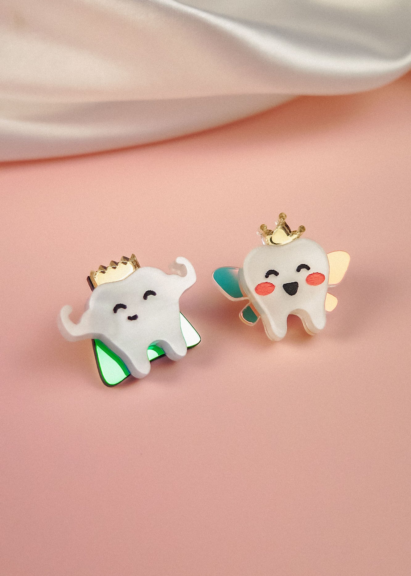 Two cute enamel pins shaped like smiling cartoon teeth, each a handcrafted accessory and true tooth king pin. One wears green wings, the other blue and gold with rosy cheeks, both crowned in gold against a soft pink background.