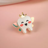 A cute Tooth Fairy Pin shaped like a smiling tooth with pink cheeks, a golden crown, and pastel wings celebrates childhood milestones, all handcrafted and set against a soft pink background.