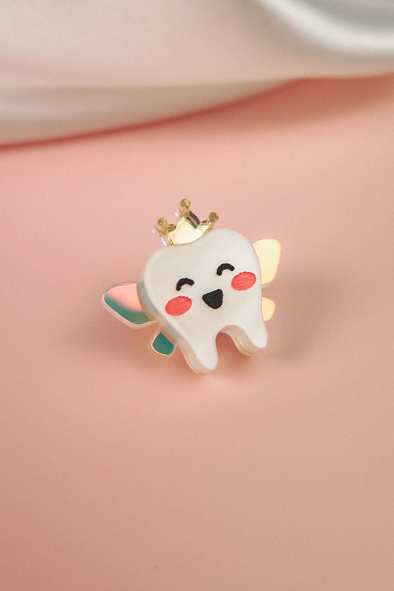 A cute Tooth Fairy Pin shaped like a smiling tooth with pink cheeks, a golden crown, and pastel wings celebrates childhood milestones, all handcrafted and set against a soft pink background.