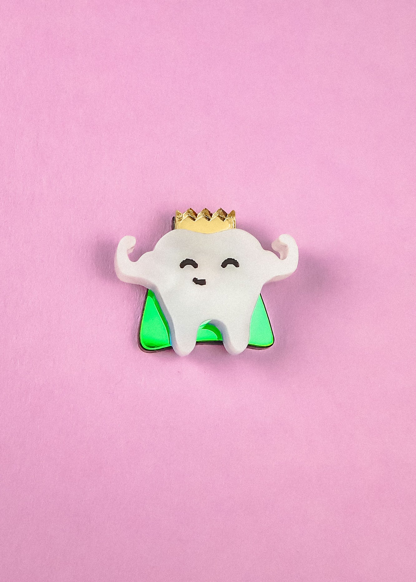A cute Tooth King Pin enamel pin features a smiling tooth with a gold crown, arms raised in front of a green triangle on a pink background. This handcrafted accessory is perfect for adding royal flair to your collection.