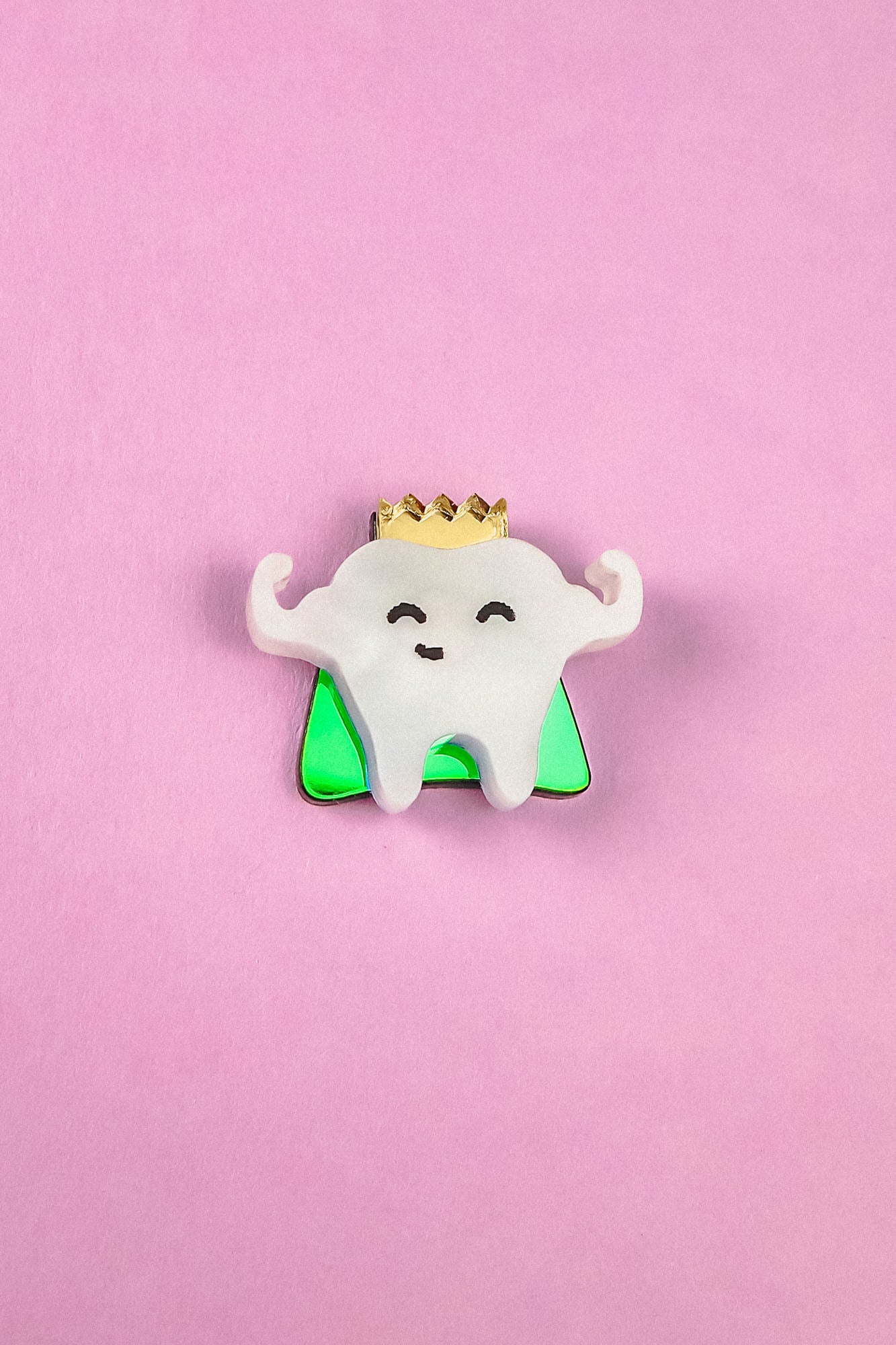 A cute Tooth King Pin enamel pin features a smiling tooth with a gold crown, arms raised in front of a green triangle on a pink background. This handcrafted accessory is perfect for adding royal flair to your collection.